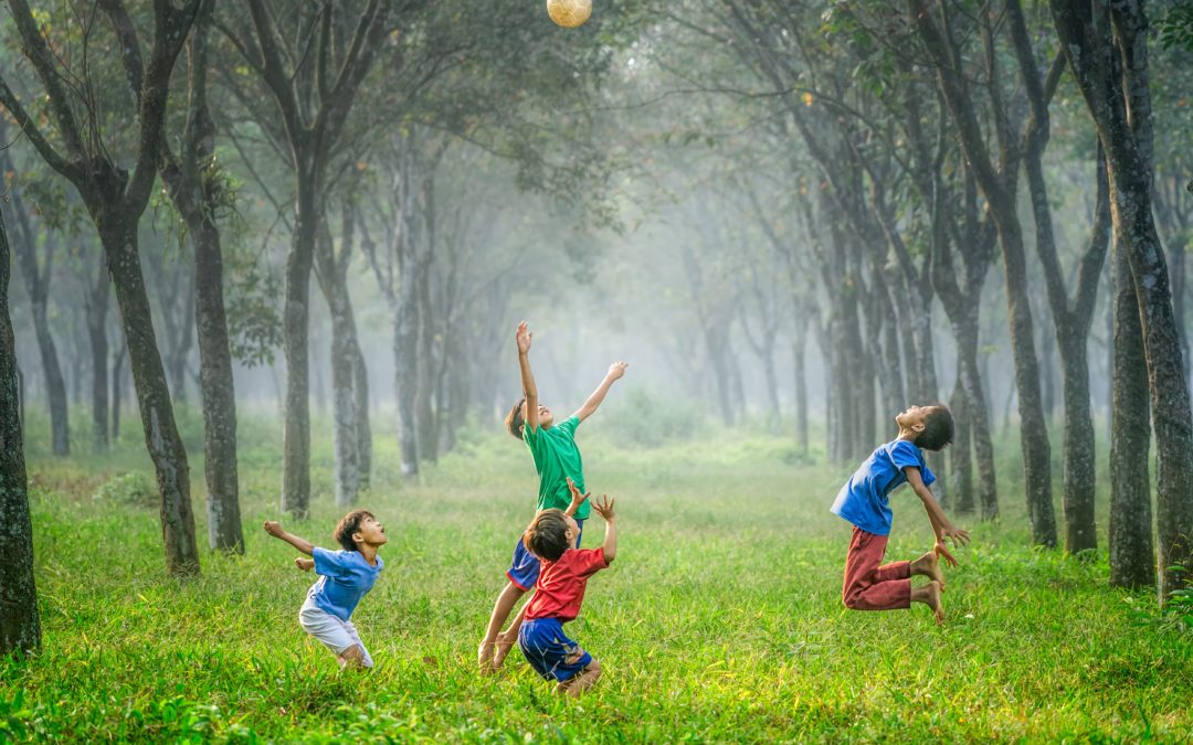 Children in bright colored clothing jumping for a ball in a grassy area surrounded by trees.
